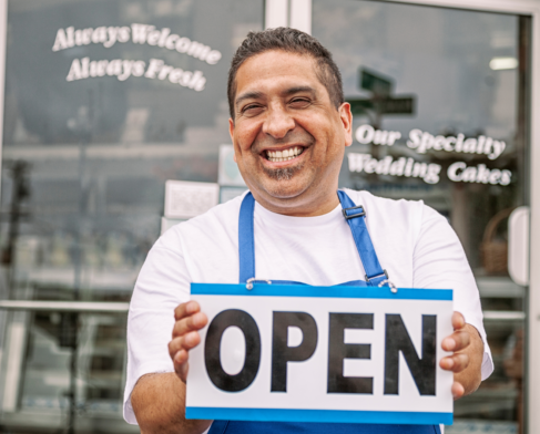 A smiling man holds an "OPEN" sign in front of the doors of a small business.