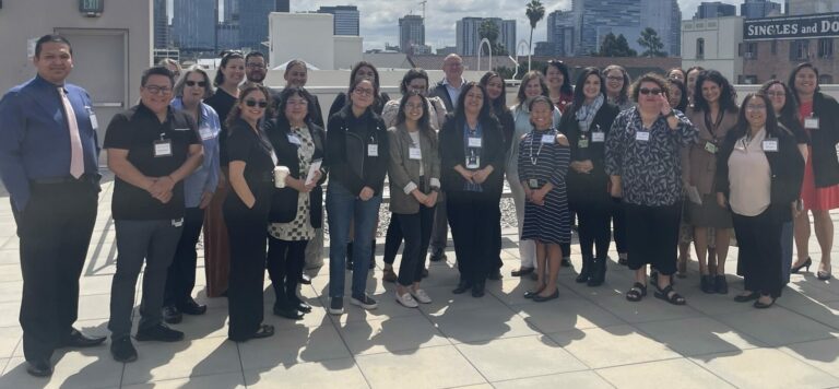 LAFLA's Medical-Legal Partnerships team and partners (about 30 people) stand together smiling on LAFLA's rooftop, with a view of cloudy Downtown LA behind them.