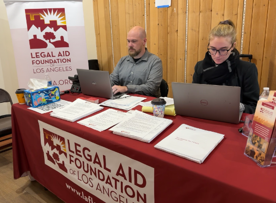 Two people sit at a table with a red tablecloth and banner both reading Legal Aid Foundation of Los Angeles. On the table are stacks of FAQs and service brochures.