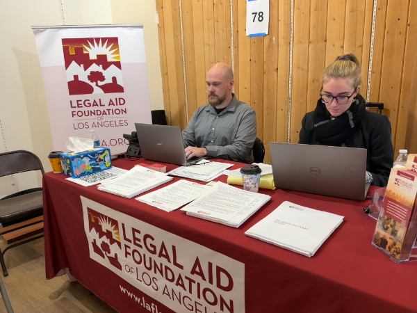 Two people sit at a table with a red tablecloth and banner both reading Legal Aid Foundation of Los Angeles. On the table are stacks of FAQs and service brochures.